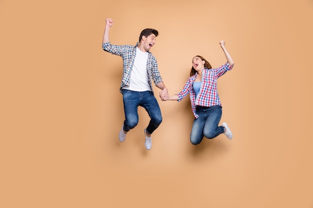 Full size photo of two people crazy lady guy jumping high celebrating best win raising fists sale shopping holding hands wear casual plaid jeans clothes isolated beige background