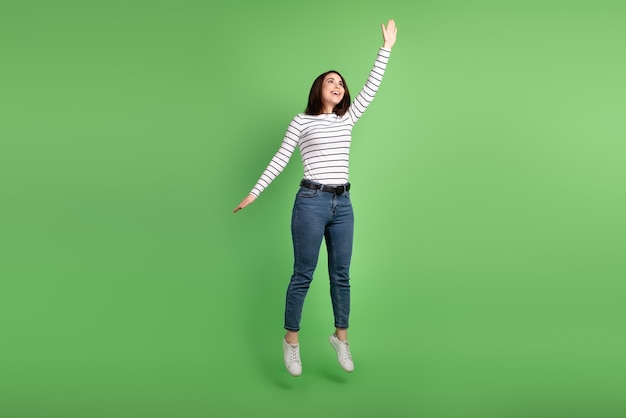 La foto a grandezza naturale di una giovane ragazza carina salta in alto e cerca di catturare l'ombrello isolato su uno sfondo di colore verde.