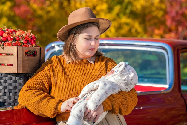 A fullbodied mother with a newborn little daughter stands near a red retro car loaded with boxes of vegetables against the backdrop of rural autumn nature