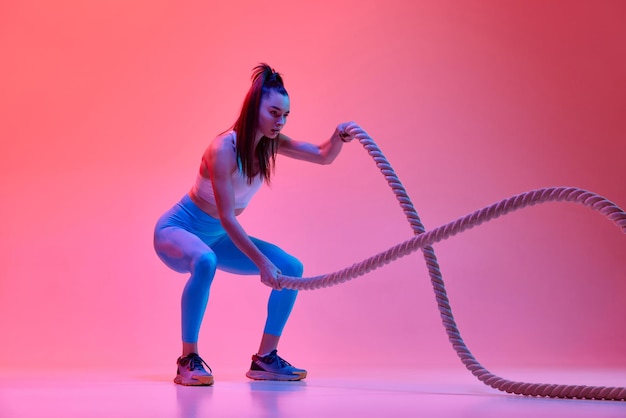 Fullbody workout Fitness Jong fit meisje training doen oefeningen met touw over roze studio achtergrond in neonlicht Concept van sport, gezondheid en kracht