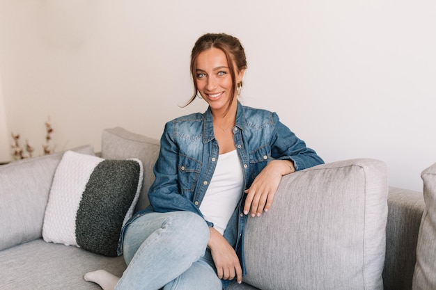 Foto a figura intera di una bella donna sorridente con i capelli scuri e un sorriso meraviglioso che indossa una camicia di jeans seduta sul divano e posa davanti alla telecamera in un appartamento moderno ed elegante