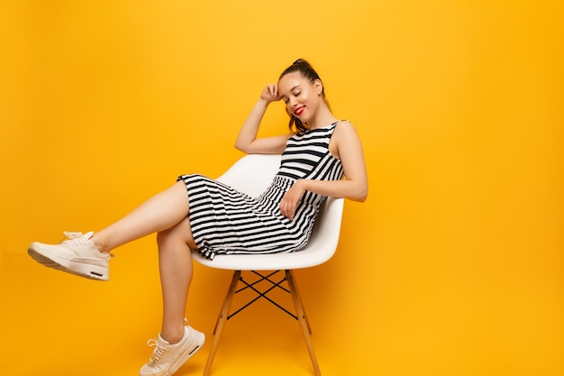 Fulllenght portrait of pretty stylish woman wearing white sneakers and stripped dress posing on yellow