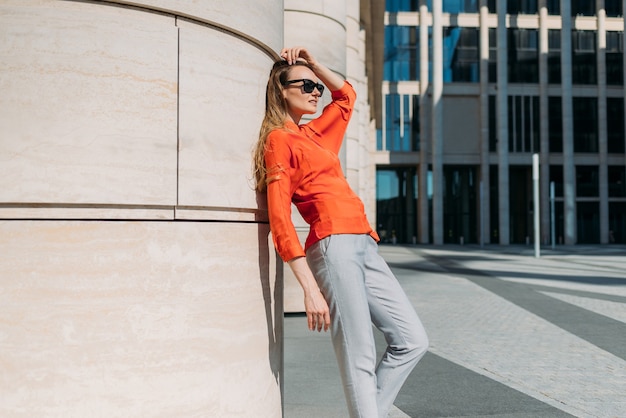 Fulllength caucasian girl in sunglasses and casual clothes leans on a building