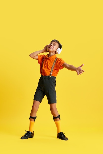 Fulllength of little boy child in stylish clothes listening to music in headphones against yellow