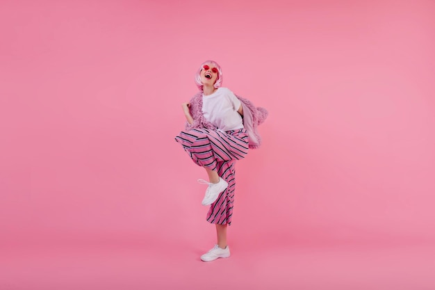 Fulllength portrait of adorable girl in bright pants dancing in studio Indoor shot of attractive female model in short periwig expressing happiness on pink background