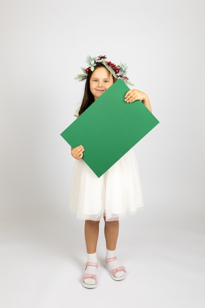 Fulllength portrait of cute girl in christmas wreath and in white festive dress holding green blank ...