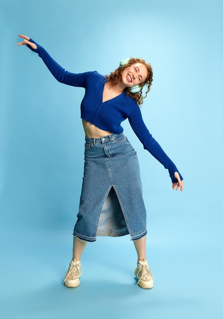 Ritratto a figura intera di giovane ragazza sorridente felice in top blu e gonna di jeans che ascolta la musica dentro