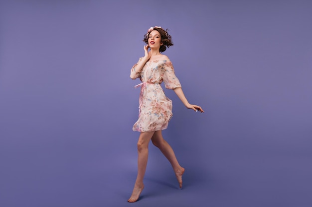 Fulllength portrait of inspired girl in summer dress jumping in studio Amazing european lady with flowers in hair dancing on violet background