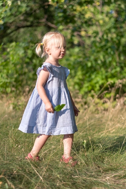 Fulllength portrait of little blonde girl in blue dress with green leaf in her hands in the park Vertical frame