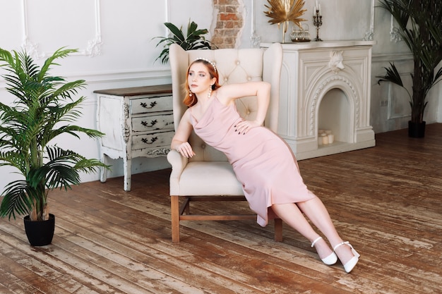 Fulllength portrait of a redhaired woman in a beige silk dress sitting in a vintage armchair on the ...