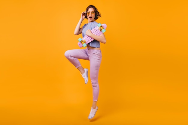 Fulllength portrait of stunning girl in yellowcolored glasses expressing amazement Indoor shot of cheerful brunette lady with longboard jumping on orange background