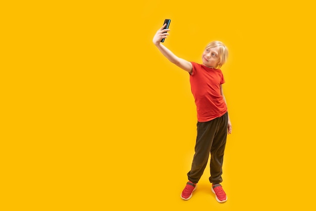 Fulllength portrait of whitehaired boy in red tshirt with phone in his hands child takes selfie isolated on yellow background copy space