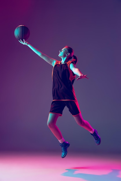 Fulllength portrait of young girl training basketball throwing ball isolated over purple background in neon