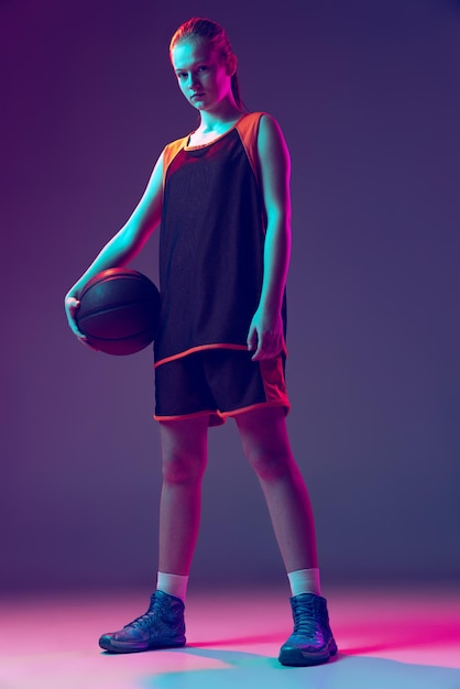 Fulllength portret van jonge sportieve meisje basketbalspeler in uniform poseren geïsoleerd over paarse achtergrond in neon