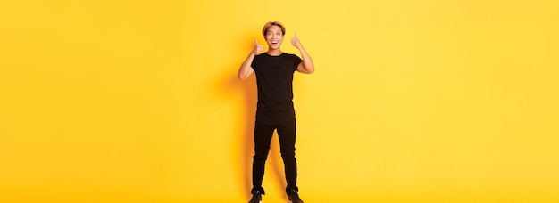 Fulllength of satisfied handsome asian guy smiling showing thumbsup in approval yellow background