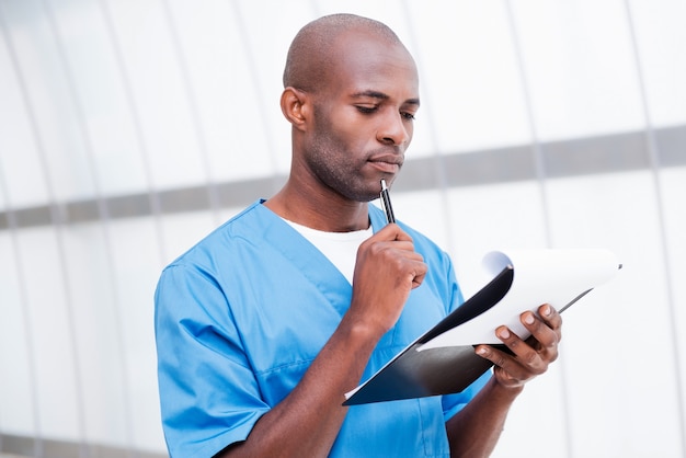 Fully concentrated at work. Thoughtful young African doctor in blue uniform holding clipboard and looking at it while touching his chin with pen