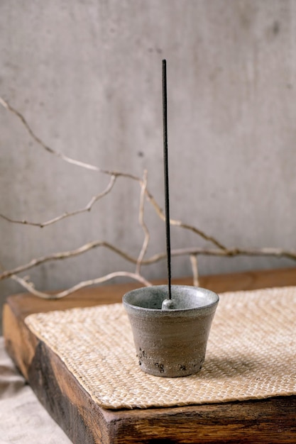 Fuming incense stick in hand crafted gray ceramic incense holder on wooden table with dry branch grey wall at background