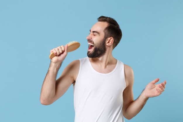 白いシャツを着た20代の楽しいひげを生やした男は、青いパステルカラーの壁、肖像画に分離されたマイクのようなホールドコームを歌います。スキンケアヘルスケア美容手順の概念。