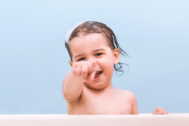 Fun cheerful happy toddler baby taking a bath playing with foam Little child in a bathtub points with his finger Smiling kid in bathroom on blue background Toddler washing and bathing Health care