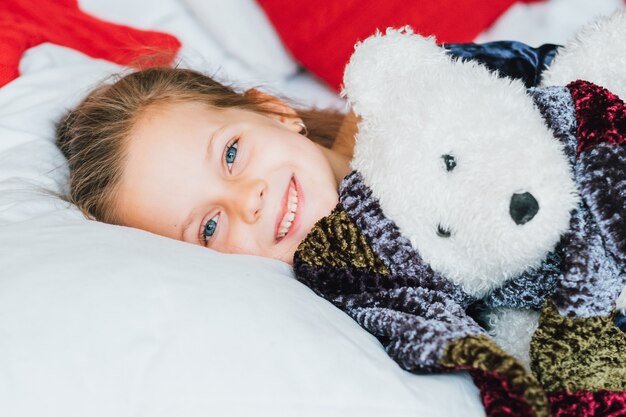 Fun childhood holidays. Cute little girl woke up in bed with teddy bear gift, smiling.