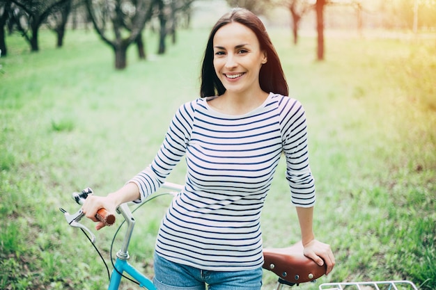 Веселый день с велосипедом Довольно великолепная и счастливая женщина со старинным велосипедом в городском парке на закате