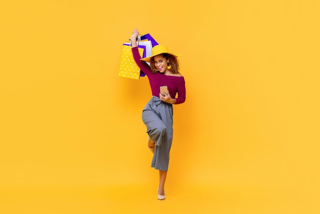 Fun full length portrait of happy young attractive shopaholic African American woman raising shopping bags and holding mobile phone in isolated studio yellow background