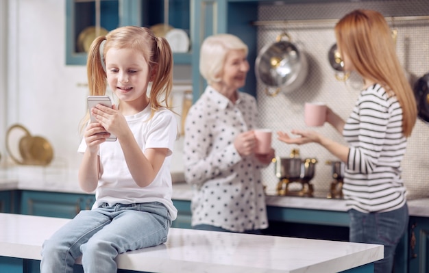 Fun game. adorable little girl sitting on the kitchen counter and playing on her phone while her mother and grandmother chatting and drinking coffee in the background