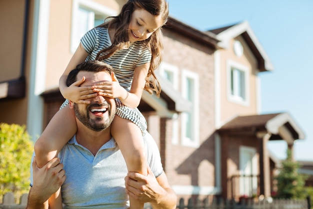 Fun game. Cute little daughter sitting on her the shoulders of her happy father and covering his eyes with her hands