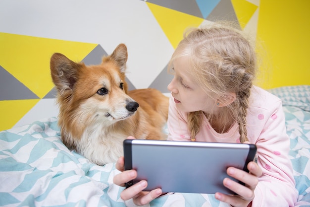 Fun girl and dog corgi on the bed in the nursery play on the tablet