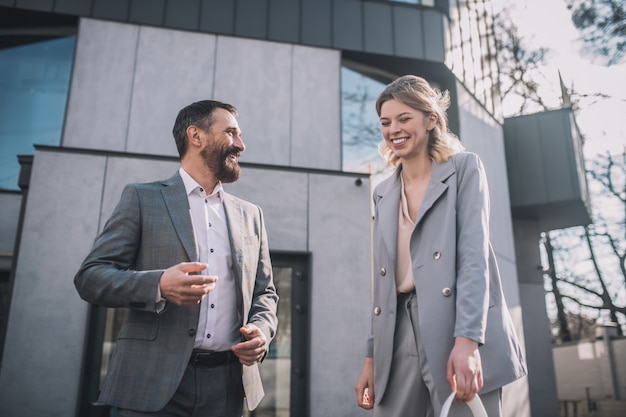 Fun mood. Business adult bearded man talking and young blonde woman laughing communicating standing outdoors near office