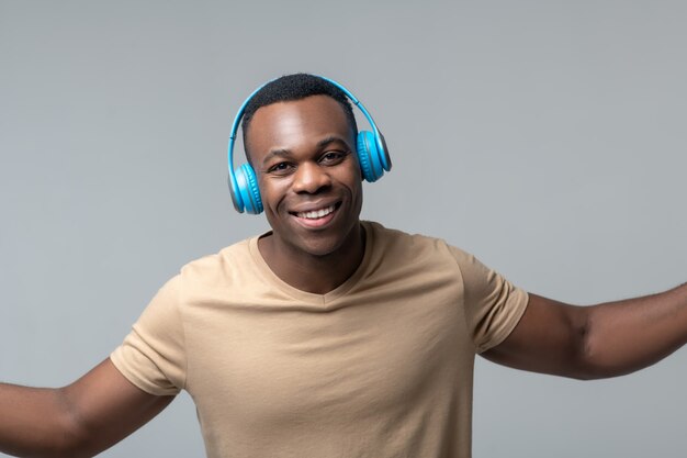 Fun mood. Joyful young adult african american in wireless headphones standing holding hands to sides in studio