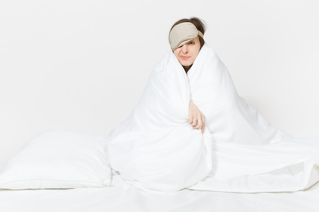 Fun tired young woman sitting in bed with sleep mask, sheet, pillow, wrapping in blanket isolated on white wall