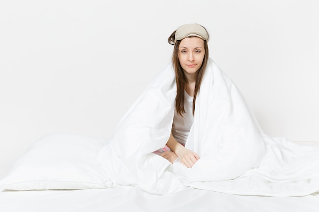 Photo fun tired young woman sitting in bed with sleep mask, sheet, pillow, wrapping in blanket isolated on white wall