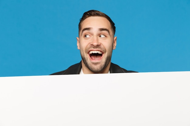 Il giovane uomo barbuto divertente guarda fuori il grande tabellone per le affissioni in bianco vuoto bianco per il contenuto promozionale isolato sul ritratto dello studio del fondo blu della parete. concetto di stile di vita di persone sincere emozioni. mock up copia spazio.