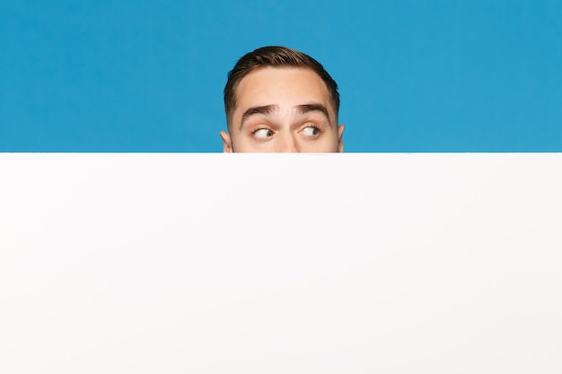 Фото Забавный молодой человек прячется, смотрит глаза большой белый пустой пустой рекламный щит для рекламного контента, изолированных на синем стенном фоне студийного портрета. концепция образа жизни эмоции людей. копируйте пространство для копирования.