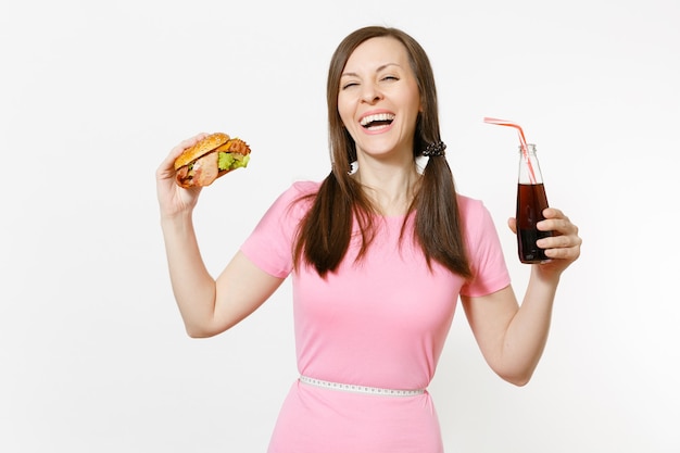 Fun young woman with tails, measuring tape at waist holding burger, cola in glass bottle isolated on white background. Proper nutrition or American classic fast food. Advertising area with copy space.