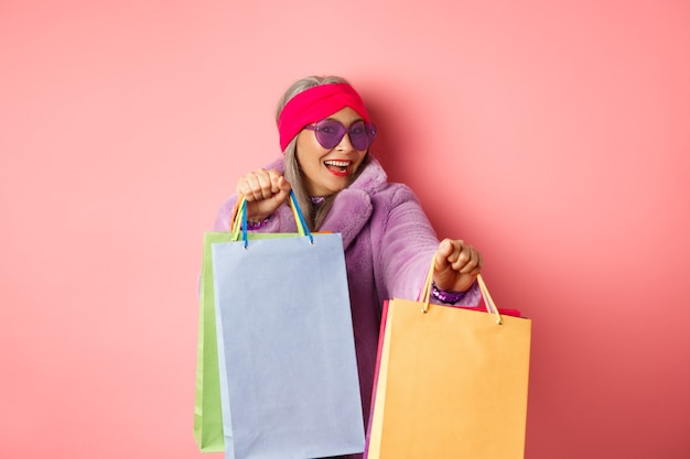 Funky and cool asian senior woman in fashionable clothes dancing while going shopping on sales, holding shop paper bags and having fun, pink background