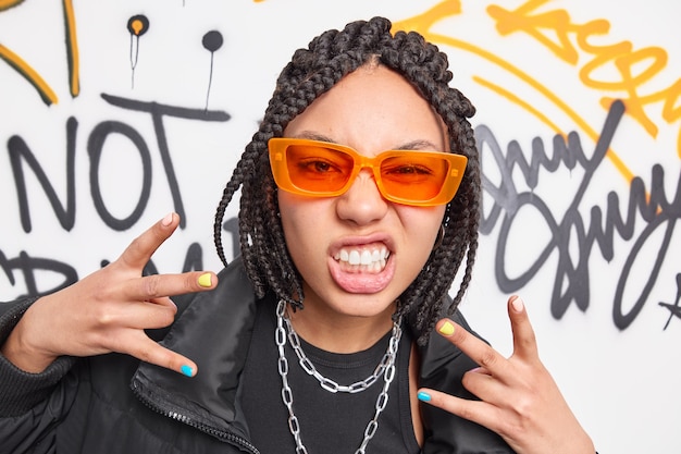 Photo funky ethnic teenage girl clenches teeth makes yo gesture feels cool has dreadlocks hairstyle wears orange sunglasses black jacket and metal chains around neck stands against graffiti wall