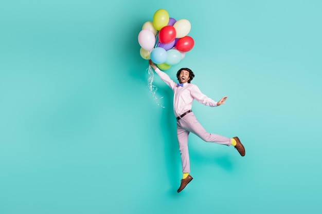 Funky jumping guy hold colorful helium balloons rise up air\
wear formal outfit isolated on turquoise background