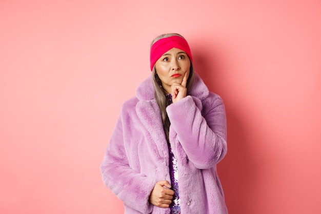 Funky old asian woman looking thoughtful and perplexed, standing in fake fur coat and thinking, making decision, standing on pink background.