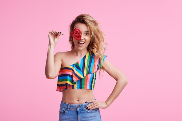 Funky positive cheerful attractive lady with her stylish trendy wave curly hair, holds a sugar sucker on stick isolated on pink