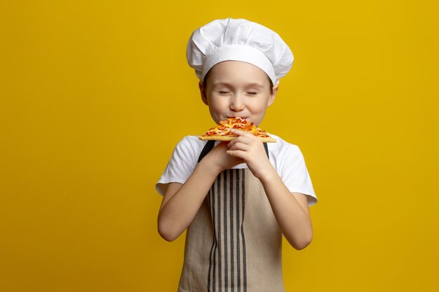 Funny 6-7 year old european-caucasian boy chef eating pizza kid in a chefs hat and a beige apron person