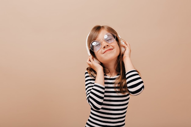 Funny adorable little girl in glasses and stripped dress enjoying music in headphones with smile and closed eyes