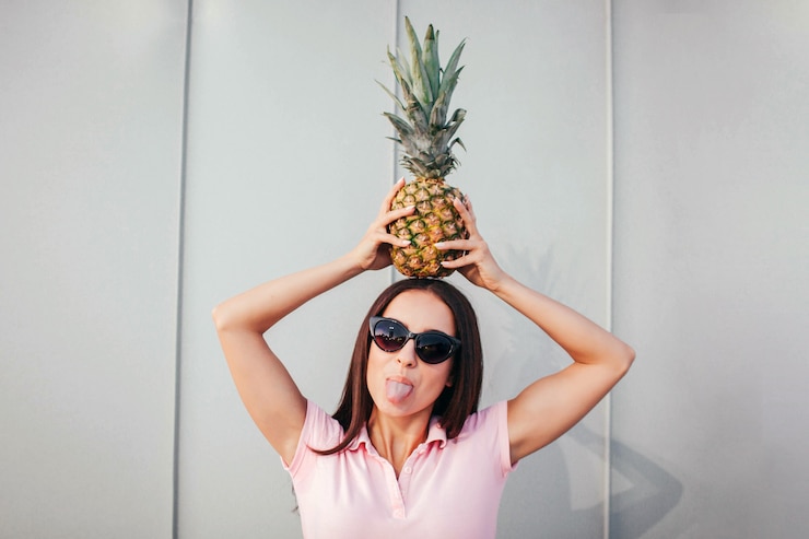 Photo funny adult girl stands and holds pineapple on her head. she wears sunglasses. woman shows her tongue.