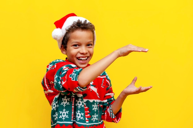 Funny african american boy in christmas clothes holds empty hands in his hands on yellow background