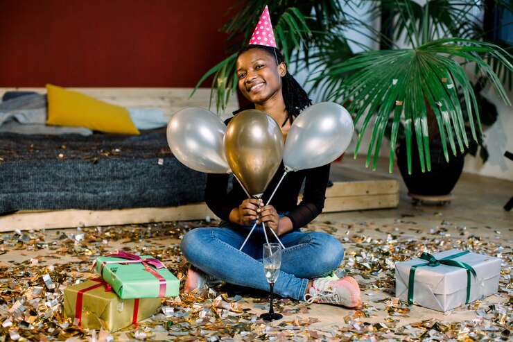 Photo funny african american girl in birthday hat and with air balloons sitting on the floor with present boxes gifts and confetti. celebration, party concept