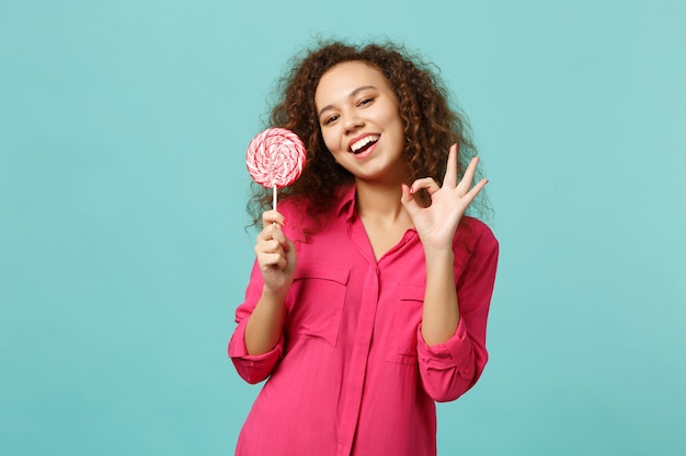 Funny african girl in casual clothes hold in hand pink round lollipop showing OK gesture isolated on blue turquoise background in studio. People sincere emotions lifestyle concept. Mock up copy space.