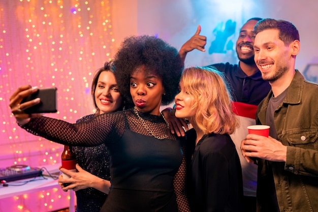 Funny African girl with wavy hair and her joyful friends looking at smartphone camera while making selfie at home party