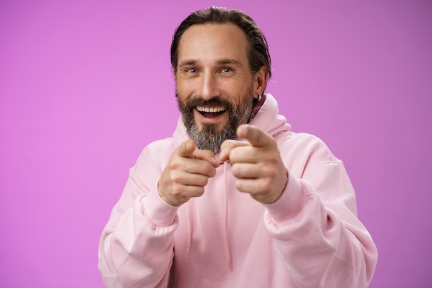 Funny amused carefree happy adult handsome man pranking friend fool around pointing camera index fingers greeting choosing you laughing joyfully having fu, standing purple background rejoicing.