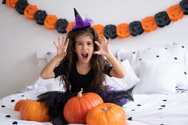 Funny arabic kid girl in witch costume having fun in decorated for halloween bedroom.